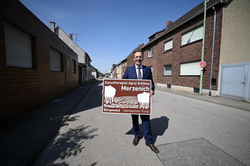 <p>Der Bürgermeister der Gemeinde Niederzier Georg Gelhausen steht mit einem entwurf für ein Hinweisschild auf einer Straße.</p>