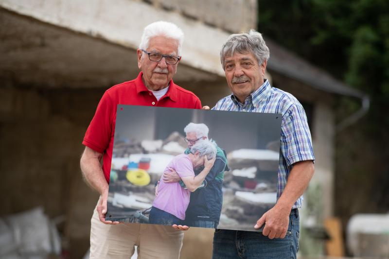 <p>Die Brüder Gerd und Bernd Gasper (rechts) stehen am ehemaligen Standort ihres bei der Flut zerstörten Elternhauses in Altenahr-Altenstadt. Dabei halten sie ein dpa-Foto in den Händen, auf dem sie sich nach der tödlichen Flutwelle vor einem Jahr weinend in den Armen liegen.</p>